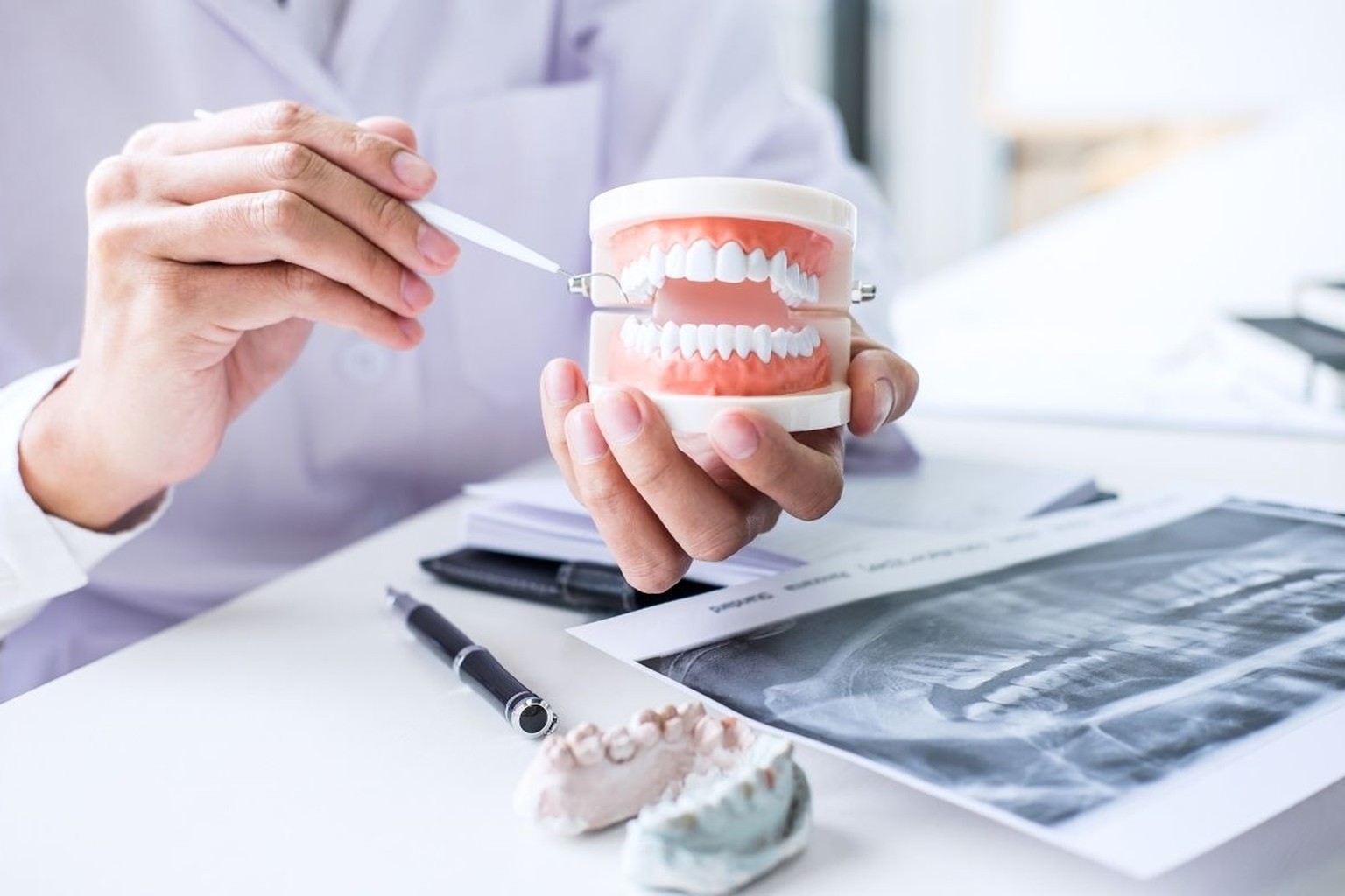 Mani in camice indicano con una sonda un modello di dentatura su scrivania bianca, accanto a calchi in gesso e radiografia