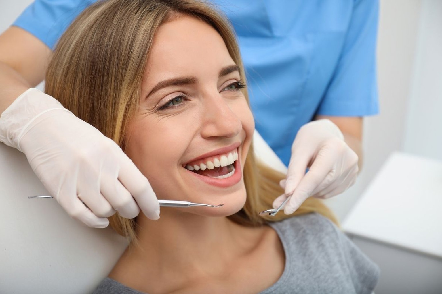 Donna bionda sorridente sulla poltrona, mani in guanti bianchi con specchietto e sonda del dentista in camice azzurro